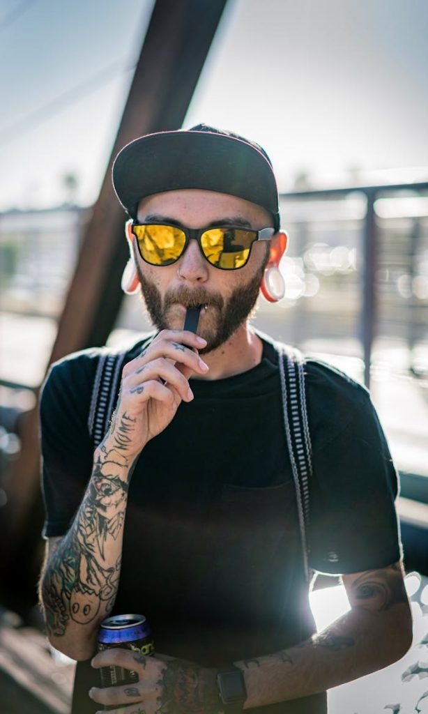 Stylish tattooed man with sunglasses vaping outdoors. Captures casual lifestyle vibe.